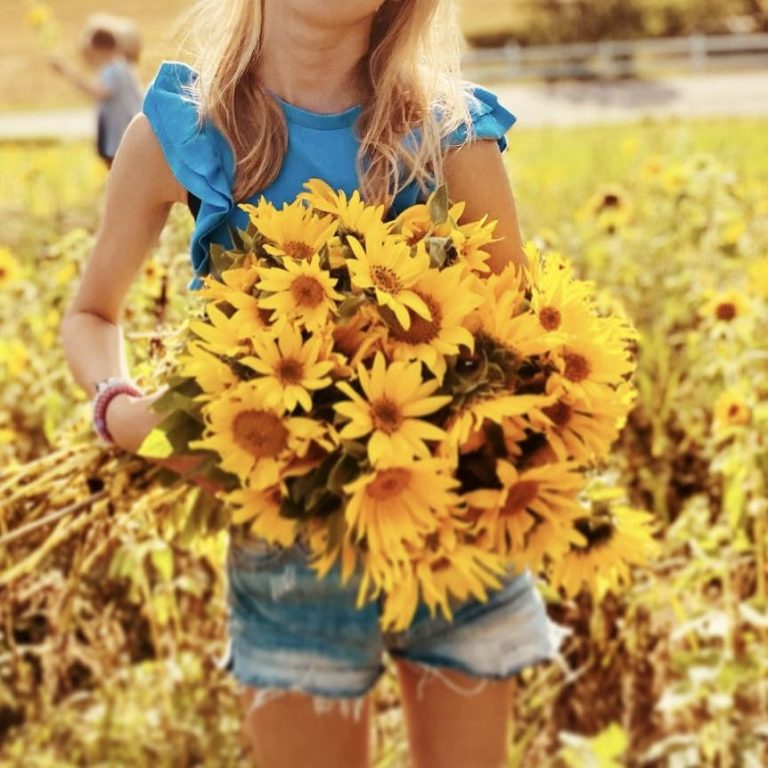 Om mig Kvinna med blont hår i röd tröja, ler framför rosa blommor och grönska.
